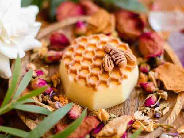 Heel and Elbow Lotion Bar is a solid skincare product. It looks like a honeycomb with a honey bee that sits on top of it. The lotion bar sists on a wooden plate surrounded by rose petals