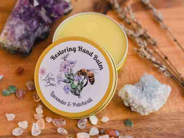 Ninas Bees Restoring Hand balm is set in a golden aluminium tin. The tin is half open, and the golden colour balm is visible in the tin. The product is placed next to crystal and lavender. Restoring Hand Balm with lavender and patchouli. Hand Balm for dry hands