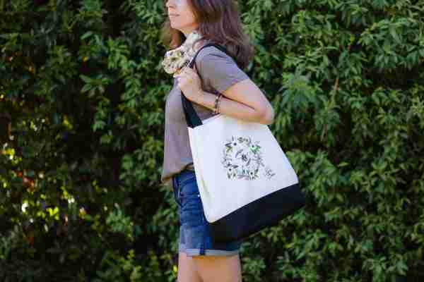 Woman holding a canvas tote bag with a floral wreath and honey bee illustration, set against a green, leafy background.