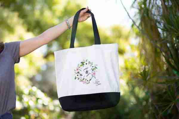 Woman holding a canvas tote bag with a floral wreath and honey bee illustration, set against a green, leafy background.