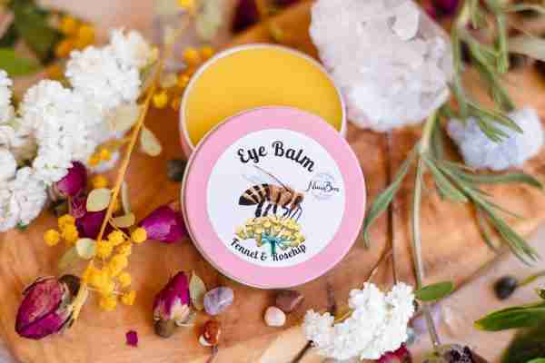 A tin of Nina's Bees Luxurious Eye Balm with fennel and rosehip, surrounded by delicate flowers and crystals on a wooden surface.