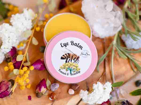 A tin of Nina's Bees Luxurious Eye Balm with fennel and rosehip, surrounded by delicate flowers and crystals on a wooden surface.