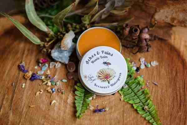Tin of Nina's Bees Aches and Pains Muscle Balm with arnica and comfrey, surrounded by natural herbs and crystals on a wooden background.