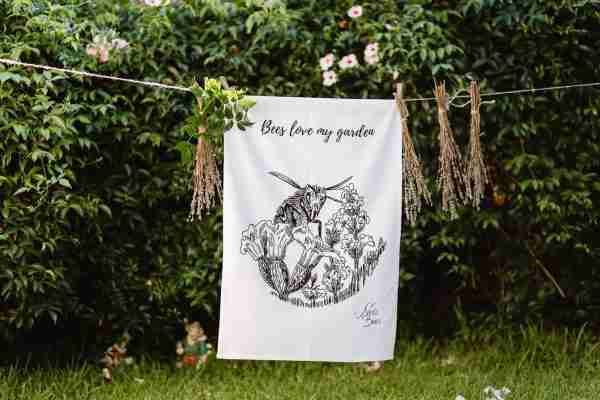 White cotton tea towel with hand-drawn bee and lavender design, “Bees Love My Garden” text, hanging on a line in a green garden.